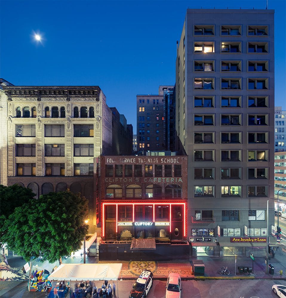 A nighttime city scene with tall buildings, a glowing neon Cliftons sign, trees, streetlights, and a cozy outdoor dining area in the foreground. The deep blue sky is illuminated by the bright moon overhead. - Hunter Kerhart